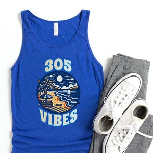 Puede incluir: Una camiseta sin mangas azul real con un gráfico de una escena de playa y el texto "305 VIBES". El gráfico está en un círculo con un esquema de color amarillo, naranja y azul. La camiseta sin mangas se combina con pantalones grises y zapatillas blancas.