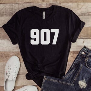 May include: Black t-shirt with the number "907" in white block letters. The shirt is paired with blue denim jeans and white canvas trainers. The items are arranged on a light brown wooden surface.