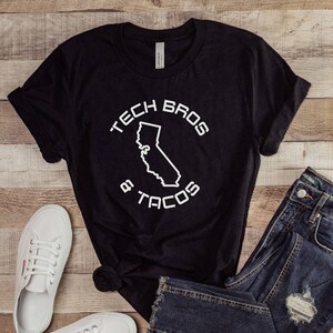 Puede incluir: Camiseta negra con el texto "TECH BROS & TACOS" rodeando un contorno blanco de California. El conjunto incluye vaqueros azules y zapatillas blancas, todo sobre una superficie de madera.