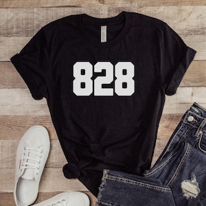 Può includere: Maglietta nera con testo bianco che dice "828". La maglietta è appoggiata su una superficie di legno con un paio di scarpe da ginnastica bianche e un paio di jeans blu.