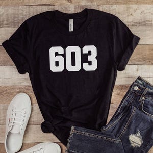 May include: Black t-shirt with the number "603" in white block letters. The shirt is paired with blue denim jeans and white canvas sneakers. The items are arranged on a wooden surface.