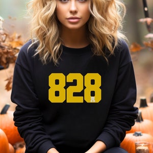 Pode incluir: Camisola preta com o número "828" em letras douradas. A pessoa usa a camisola e está sentada em frente a abóboras e folhas de outono. O fundo inclui velas e uma cena florestal desfocada.
