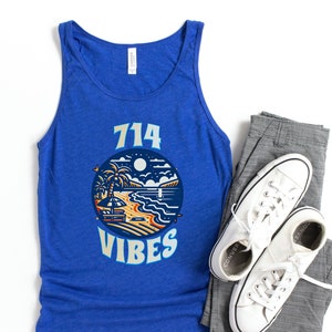 May include: A blue tank top with a graphic of a beach scene and the text "714 VIBES". The tank top is paired with gray pants and white sneakers.