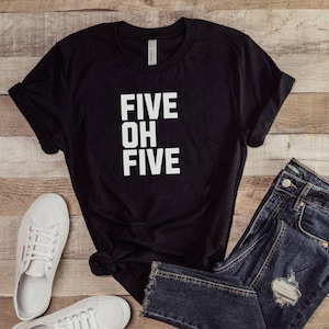 May include: Black t-shirt with the text "FIVE OH FIVE" in white, paired with blue jeans and white sneakers. The shirt is knotted at the waist, and the jeans have a distressed patch. The items are arranged on a wooden surface.