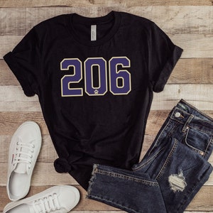 Könnte beinhalten: Schwarzes T-Shirt mit einer großen, in Lila und Gold gedruckten Nummer 206 auf der Vorderseite. Das Shirt wird mit einer blauen Jeans und weißen Turnschuhen kombiniert.