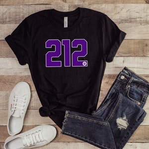 Puede incluir: Camiseta negra con el número "212" en morado impreso en la parte delantera. La camiseta se combina con unos vaqueros azules y zapatillas blancas.
