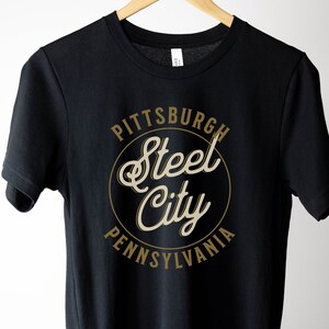 May include: Black t-shirt with a gold circle graphic that says "Pittsburgh Steel City Pennsylvania".