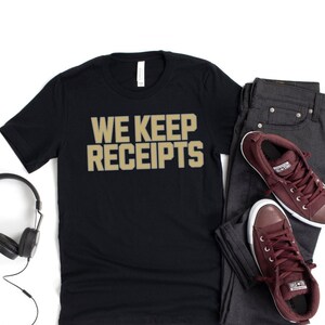 May include: A black t-shirt with the text "WE KEEP RECEIPTS" in gold lettering. The shirt is paired with dark gray jeans and maroon sneakers.