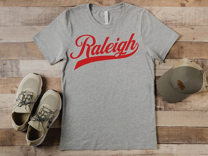 May include: A gray t-shirt with a red "Raleigh" graphic in a baseball-style font. The shirt is laid flat on a wooden surface with a brown baseball cap and two beige sneakers.