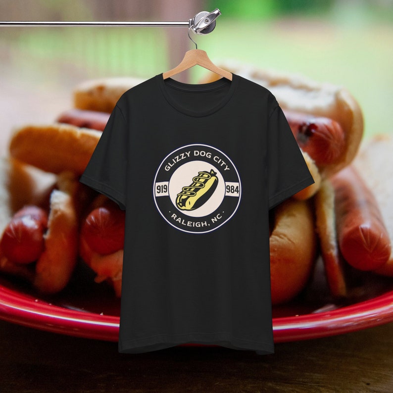 Puede incluir: Camiseta negra con un gr&aacute;fico circular que presenta un perrito caliente y el texto "Glizzy Dog City 919 984 Raleigh, NC". La camiseta est&aacute; colgada en una percha de madera sobre un fondo borroso de perritos calientes.