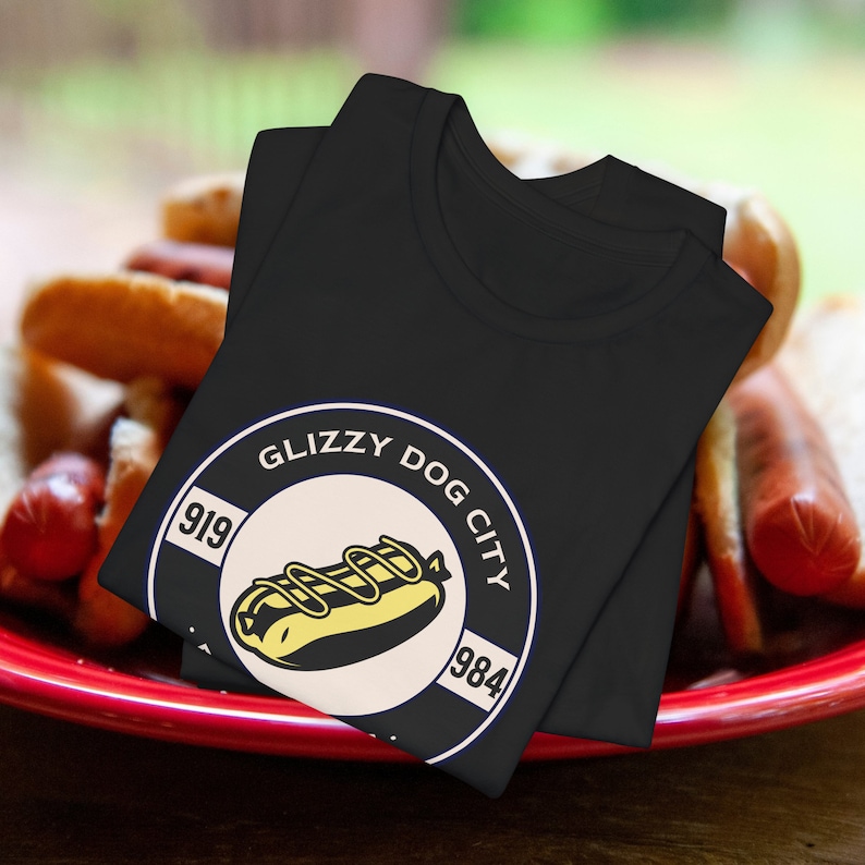 Puede incluir: Camiseta negra con un gr&aacute;fico circular que muestra un perrito caliente con mostaza y el texto "Glizzy Dog City". El gr&aacute;fico incluye los n&uacute;meros "919" y "1984". La camiseta est&aacute; doblada sobre un plato rojo con perritos calientes y panecillos.
