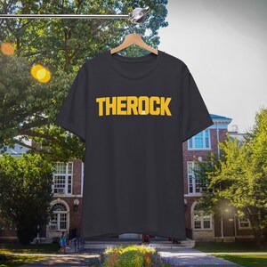 Puede incluir: Una camiseta negra con la palabra "THEROCK" impresa en letras amarillas. La camiseta est&aacute; colgada de una percha de madera. El fondo es una imagen borrosa de un edificio y &aacute;rboles.