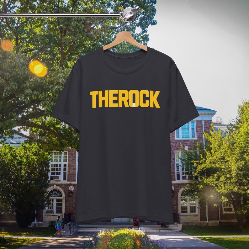 Puede incluir: Una camiseta negra con la palabra "THEROCK" impresa en letras amarillas. La camiseta est&aacute; colgada de una percha de madera. El fondo es una imagen borrosa de un edificio y &aacute;rboles.