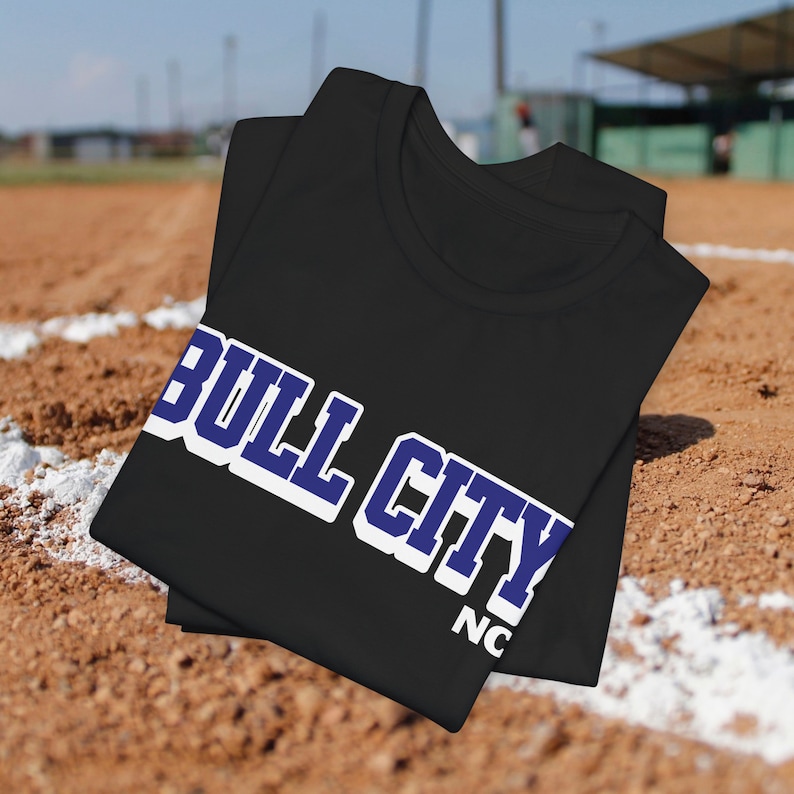 May include: Black t-shirt with the text "BULL CITY" in white and blue lettering. The shirt is folded and resting on a baseball field. The text "NC" is also visible.