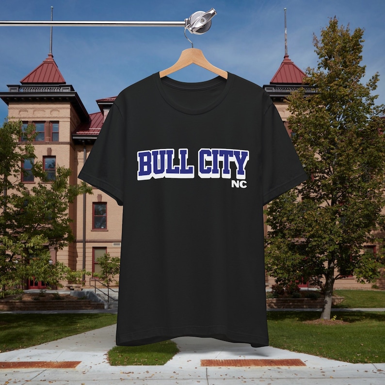 May include: Black t-shirt with the text "BULL CITY" in white and blue lettering. Below the text is "NC" in white lettering. The t-shirt is hanging on a wooden hanger against a backdrop of a building and trees.