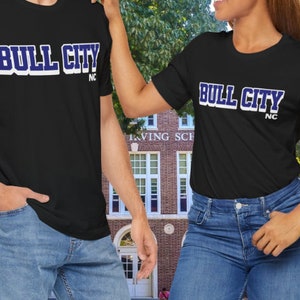 May include: Two black t-shirts with the text "BULL CITY" in white with a blue outline, and "NC" below. The shirts are worn by two people. The background is a brick building.