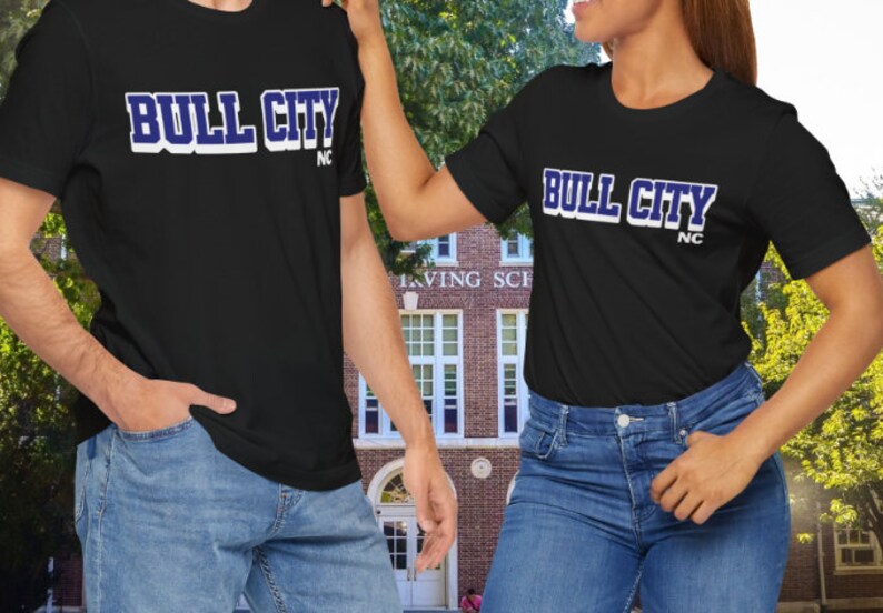 May include: Two black t-shirts with the text "BULL CITY" in white with a blue outline, and "NC" below. The shirts are worn by two people. The background is a brick building.