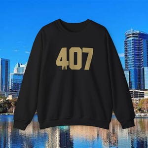 May include: Black crewneck sweatshirt with the number "407" in gold block letters across the chest. The sweatshirt has long sleeves and a ribbed neckline. The background features a cityscape with tall buildings and a body of water.
