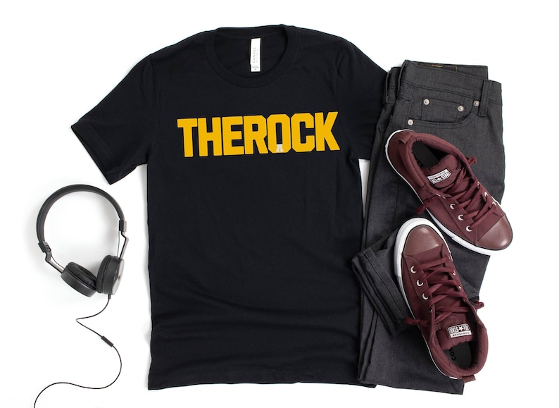 Puede incluir: Camiseta negra con la palabra "THEROCK" en amarillo. La imagen incluye vaqueros grises oscuros y zapatillas burdeos. Tambi&eacute;n se ven unos auriculares negros.