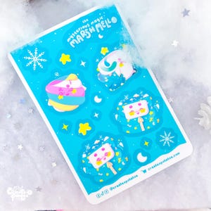Peut inclure: Planche de stickers avec des personnages de guimauve sur fond bleu clair, ornée d'étoiles et de flocons de neige. Le texte "the mysterious magic MARSHMELLO" est visible. Les autocollants ont une bordure blanche.