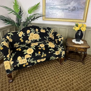 May include: Miniature black floral sofa with yellow flowers and green leaves. The sofa has curved arms and wooden legs. A small wooden side table with a black vase of yellow roses sits next to the sofa. The room has a painting and a brown rug.