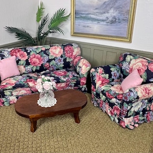 May include: A miniature dollhouse furniture set featuring a sofa and an armchair upholstered in a dark navy blue fabric with a vibrant pink, red, and white floral pattern. Each piece includes a solid light pink throw pillow and a wooden coffee table.
