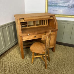 May include: A miniature wooden roll-top desk with a matching wooden chair. The desk features a curved top, a flat writing surface, a small drawer with two dark knobs, and internal cubby compartments. The chair has a simple backrest and four legs.