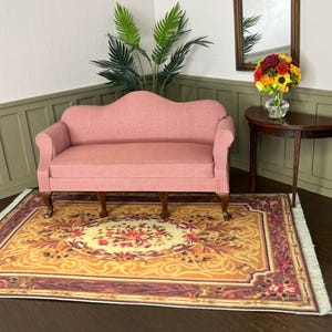 May include: A miniature pink sofa with curved arms and wooden legs is placed on a patterned rug. The rug features a central floral design in yellow, red, and brown. A small, dark wooden table holds a vase of flowers.