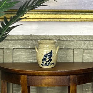 Könnte beinhalten: Ein Miniatur-Keramiktopf in Beige mit zwei kleinen Griffen und einem blauen Blumendesign. Der Topf steht auf einem Holztisch, mit einem Gemälde im Hintergrund. Der Topf ist ein dekorativer Gegenstand.