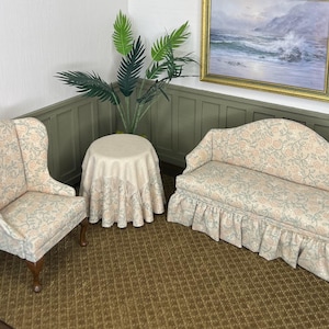 May include: Miniature living room set with a floral pattern. Includes a wingback chair, a loveseat with a skirted base, and a round table. The set is on a brown rug, with a painting and a plant in the background.