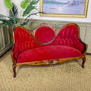May include: A miniature dollhouse sofa featuring a polished wooden frame with carved floral details and a vibrant red velvet upholstered seat and backrest. The backrest has a central circular panel and curved sides with vertical stitching.