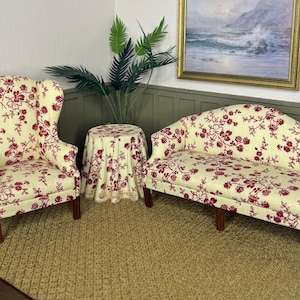 May include: Miniature living room set with a wingback chair, loveseat, and round table. The furniture features a cream-colored floral pattern with red flowers. The set is placed on a woven rug, with a painting and greenery in the background.