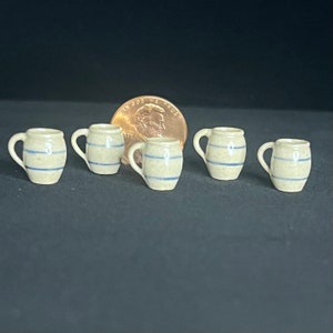 May include: Five small, cream-colored ceramic mugs with blue stripes. The mugs have a rounded body and a handle.