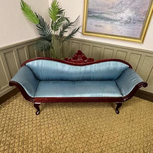 May include: A miniature antique-style sofa with a light blue velvet seat and a dark red wooden frame. The sofa has curved arms and legs, and a decorative back. The sofa is placed on a woven rug.