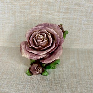 Puede incluir: Una escultura de rosa de cerámica en tonos malva y crema, con hojas verdes. La rosa grande está completamente florecida, con un capullo más pequeño en la base. La escultura es un acento floral decorativo.
