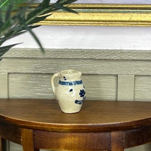 Könnte beinhalten: Ein kleiner, cremefarbener Keramikkrug mit Henkel, verziert mit blauen Blumen- und Streifenmustern. Der Miniaturkrug steht auf einem Holztisch und zeigt seinen rustikalen Charme und seine filigranen Details. Der Krug ist ein dekorativer Gegenstand.