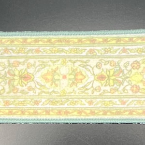 May include: A rectangular rug with a cream-colored background and a floral pattern in yellow, peach, and green. The rug has a light blue border and fringe on both ends. The rug is approximately 36 inches long.