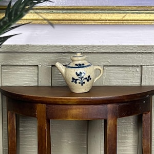 May include: A small, cream-colored teapot with blue floral designs sits on a dark brown wooden table.