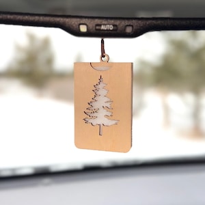 May include: A wooden car air freshener with a cut-out design of a pine tree. The air freshener is hanging from the rear-view mirror of a car.