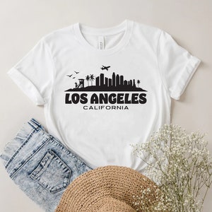 May include: White t-shirt featuring a black silhouette of the Los Angeles skyline, with the text "LOS ANGELES CALIFORNIA" in bold, black letters. The shirt is paired with blue denim shorts and a straw hat.