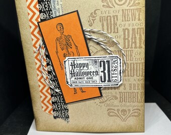May include: A Halloween-themed card with a skeleton, a ticket that says "Happy Halloween, Admit One, Good Date Sold Only", and the number 31. The card has a vintage, distressed look with a brown background and orange, black, and white accents.