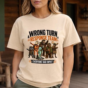 May include: A beige short-sleeve t-shirt featuring a graphic of a raccoon, a masked figure, an alien, a sasquatch, a bear, and a moose. The text reads WRONG TURN RESPONSE TEAM and EVERYONE HAD INPUT in black and orange lettering.