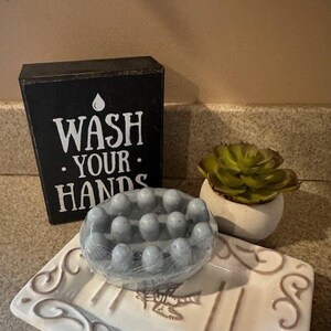 May include: A gray soap bar with a textured surface sits on a white ceramic soap dish with a decorative design. The soap dish is on a countertop next to a black wooden sign that reads "Wash Your Hands" with a water drop icon. A small potted succulent plant is also on the countertop.