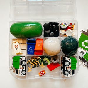 May include: A clear plastic storage box filled with various items for a pretend play garbage truck theme. The box contains green and white play dough, black stones, a small toy garbage truck, a toy construction barrier, a toy recycling bin, and other small toys.