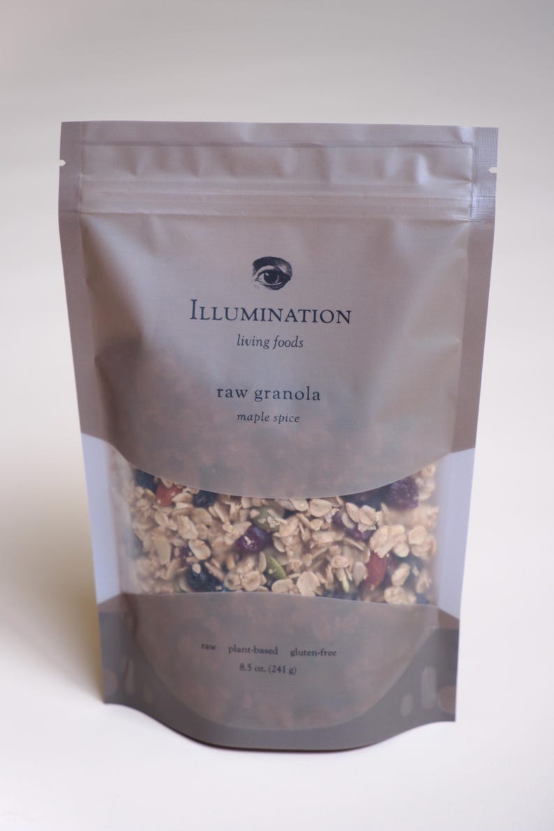 Puede incluir: Un paquete de granola cruda con sabor a arce. El envase es de color marr&oacute;n claro con una ventana transparente que muestra la granola. El texto "ILLUMINATION living foods" est&aacute; impreso en la parte superior, y "raw granola maple spice" debajo. El paquete contiene 241 g de granola.