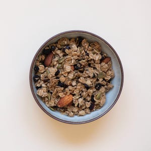 Puede incluir: Vista superior de un cuenco lleno de granola. La granola es una mezcla de avena, nueces, semillas y frutos secos. El cuenco es azul claro con un borde marr&oacute;n, sobre un fondo color crema.