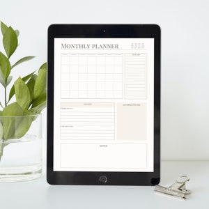 May include: A black tablet with a white monthly planner on the screen. The planner includes sections for to-do lists, goals, affirmations, and notes. The tablet is on a white surface.