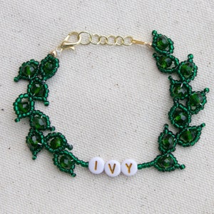 May include: A green beaded bracelet with a leaf design. The bracelet features the word "IVY" spelled out in white letter beads. The bracelet has a gold-colored clasp and extender chain.