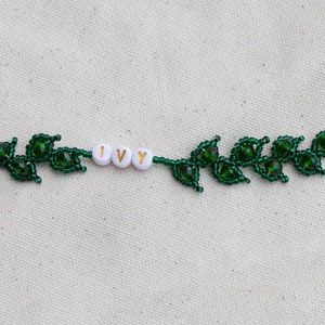 May include: A beaded bracelet featuring a green leaf design. The bracelet includes the word "IVY" spelled out in white beads. The bracelet has a gold-colored clasp and chain for adjustable sizing. The leaves are made of dark green beads.