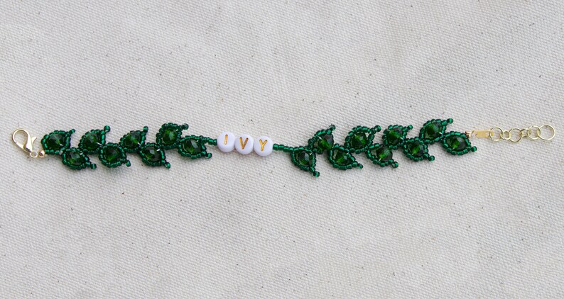 May include: A beaded bracelet featuring a green leaf design. The bracelet includes the word "IVY" spelled out in white beads. The bracelet has a gold-colored clasp and chain for adjustable sizing. The leaves are made of dark green beads.